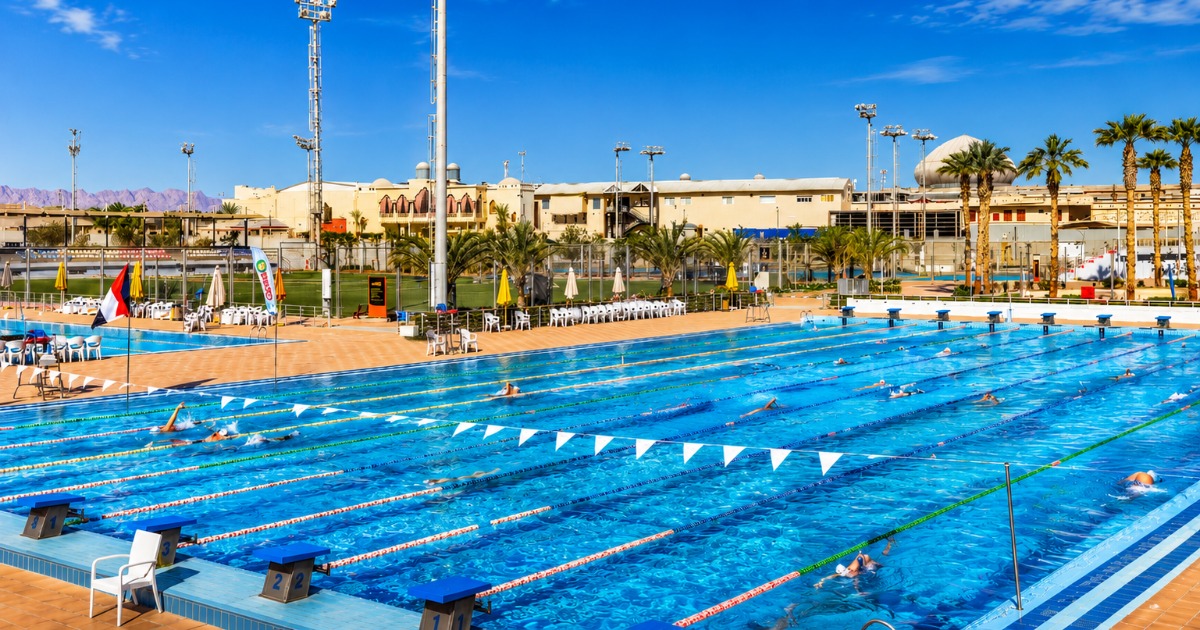 Wide view of the 50m Olympic outdoor pool in active use with swimmers training across multiple lanes, multi-sport complex buildings and floodlit pitches visible in the background, Hurghada, Egypt