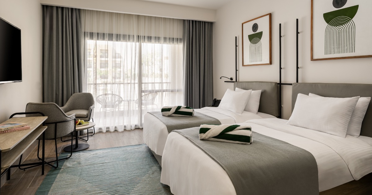 Twin room with two beds, floor-to-ceiling curtains, balcony, wall-mounted TV, and desk area at the Soma Bay resort hotel