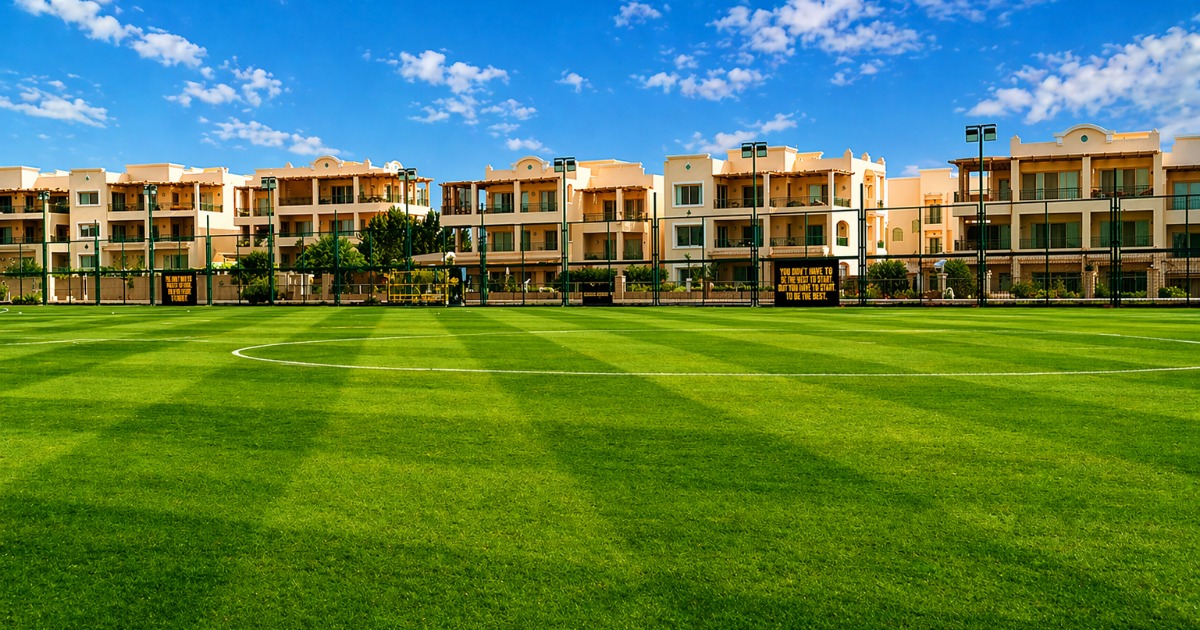 Ground-level view of a natural grass football pitch under a blue sky, with the resort's Mediterranean-style accommodation buildings directly behind the perimeter fence