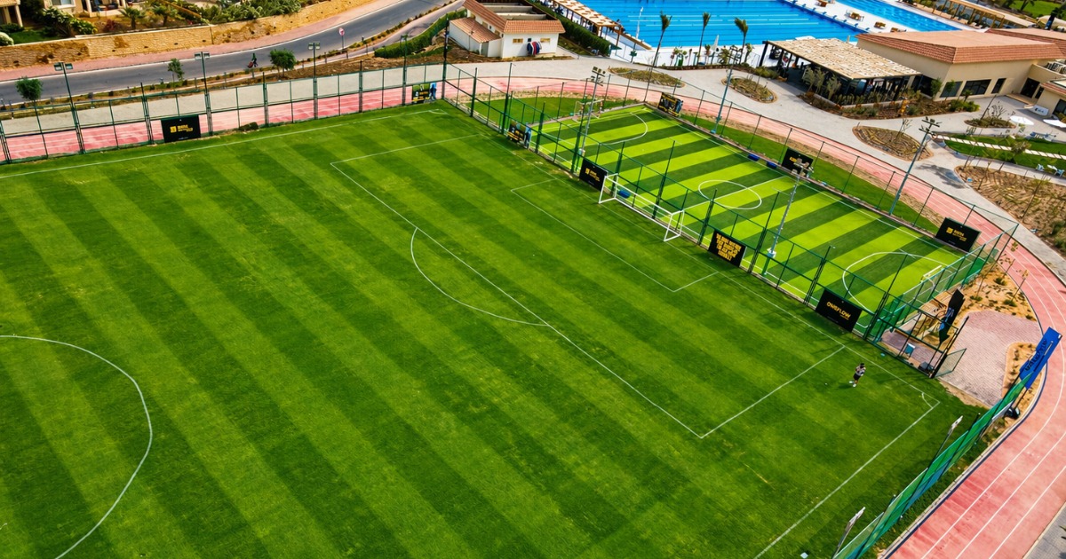 Aerial view showing a full-size grass training pitch and adjacent smaller artificial turf pitch at Soma Bay sports complex, with an outdoor swimming pool visible in the top right