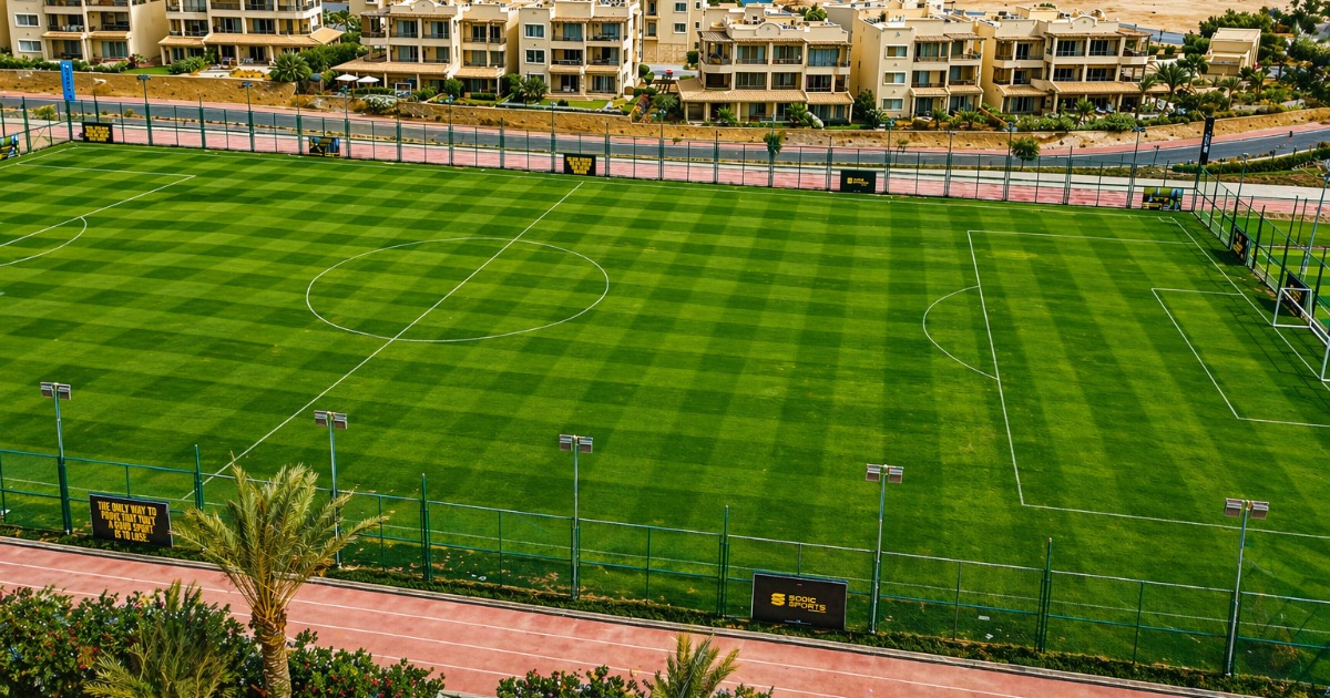 Elevated view of two natural grass football pitches at the Hurghada sports complex, with a running track surround and resort villas in the background