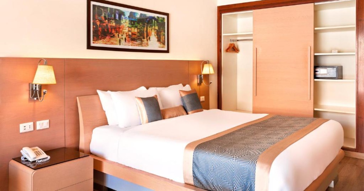 Standard double room with warm wood headboard, white bedding, and built-in wardrobe with safe