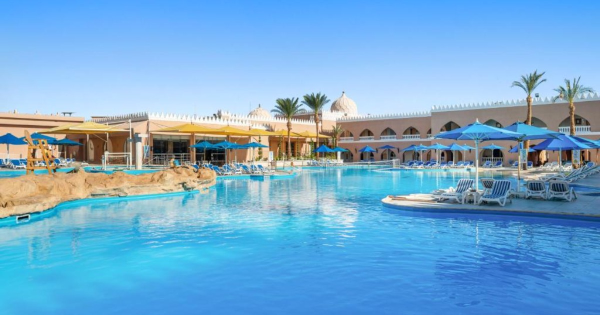 Large turquoise leisure pool with sun loungers and blue umbrellas, resort arcade and domed architecture in the background