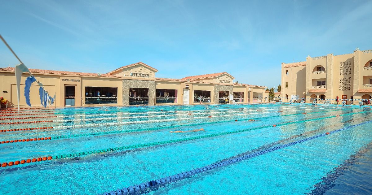 Wide view of 50m resort swimming pool with lane ropes, swimmers, and hotel buildings in Hurghada
