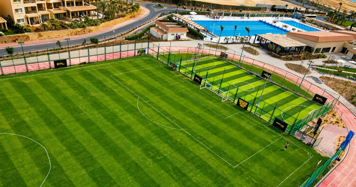Aerial view of a full-size football pitch and adjacent smaller 5-a-side court with alternating grass stripe patterns, next to a running track and outdoor swimming pool at a resort complex
