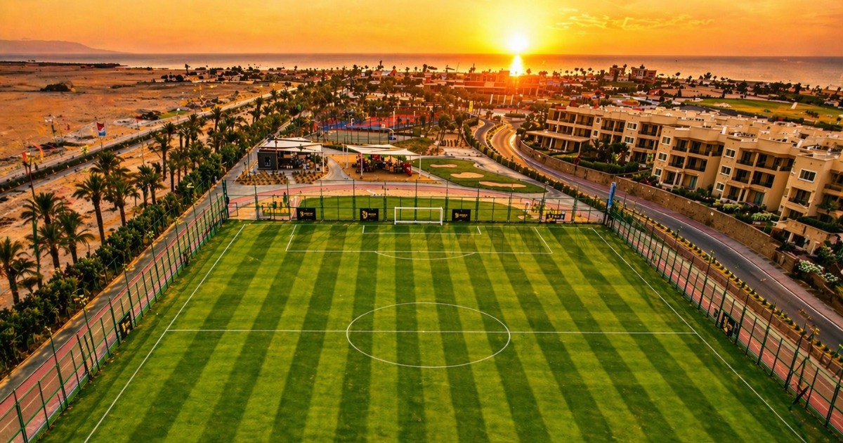 Drone aerial view of a floodlit football pitch at golden sunset with the Red Sea coastline and palm-lined resort town visible in the background