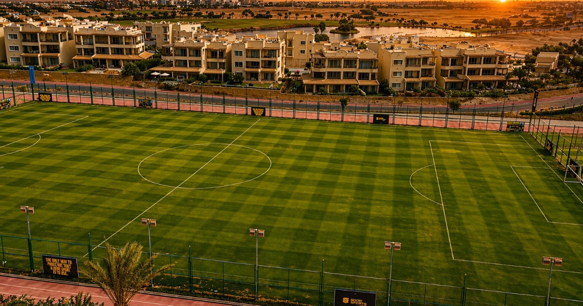 Elevated view of a full-size football pitch bathed in warm golden sunset light, with a running track perimeter, resort villas, and a tranquil lagoon visible in the background