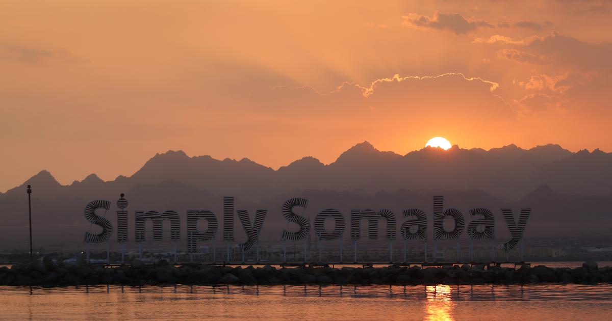 Simply Somabay large letter sign reflected in calm water at sunset, with mountain silhouette behind