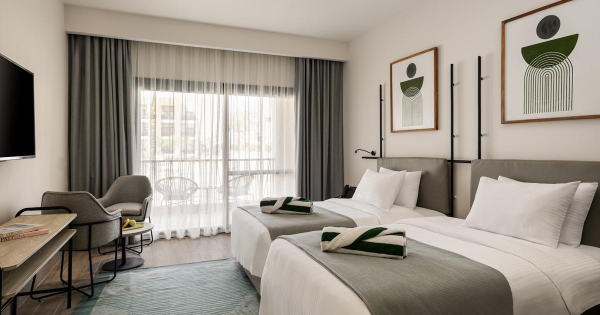 Bright twin hotel room with two beds, balcony, geometric wall art, and a desk area
