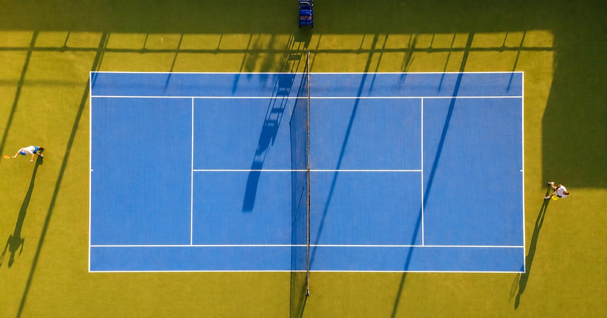 Top-down aerial view of two players in a match on a blue Soma Bay tennis court, with long shadows cast across the surface