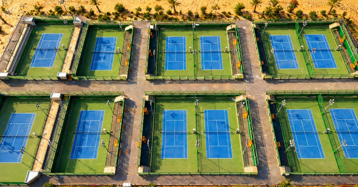 Aerial daytime view of six blue ITF tennis courts at Soma Bay arranged in two rows, surrounded by desert sand and palm trees