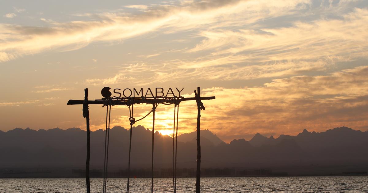 Somabay resort sign mounted on wooden poles over open Red Sea water at sunset, with layered mountain silhouettes and dramatic cloudy sky