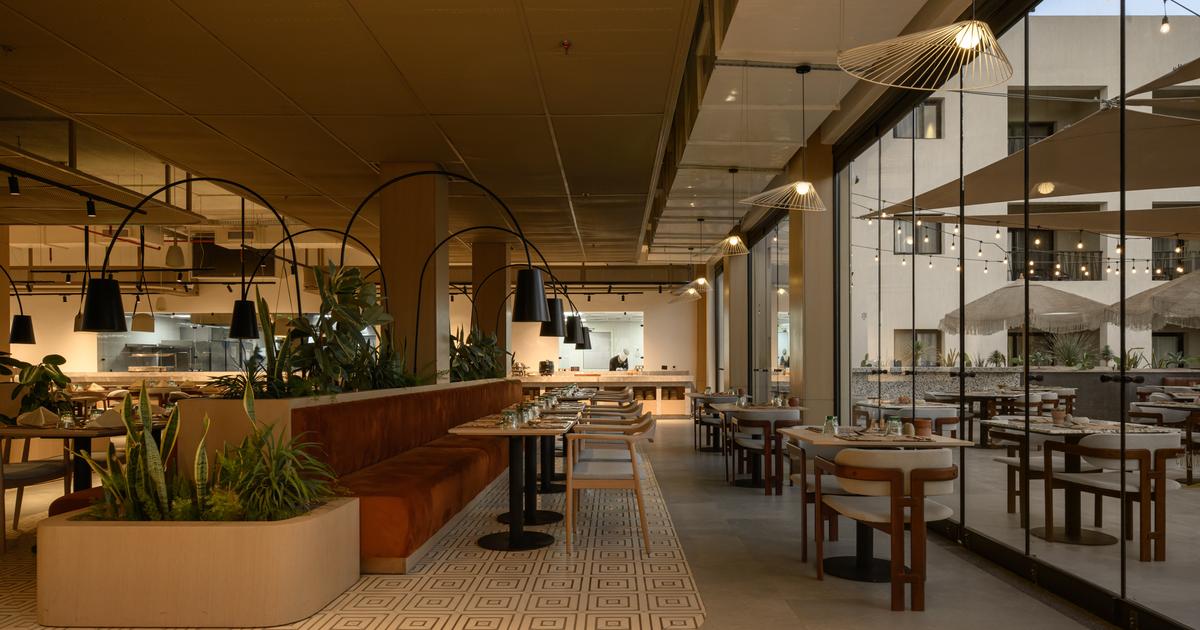 Warmly lit hotel restaurant with open kitchen, lush indoor plants, arched pendant lights, and floor-to-ceiling windows overlooking a terrace