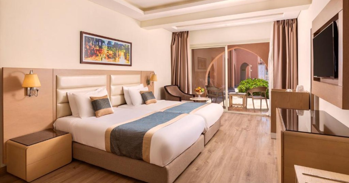 Standard hotel room at the Hurghada all-inclusive resort with double bed, warm wood furnishings, balcony with arched terrace view, and wall-mounted TV