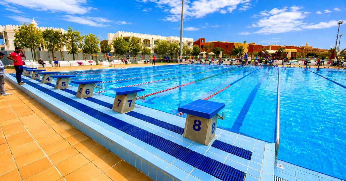 Starting blocks on lanes 3–8 of the 50m pool at Hurghada sports complex, with athletes preparing on deck and resort architecture visible in the background